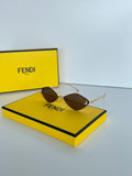 Fendi sunglasses with gold-tone metal frames and dark brown lenses, displayed on top of a bright yellow Fendi box with the brand's signature yellow box in the background.
