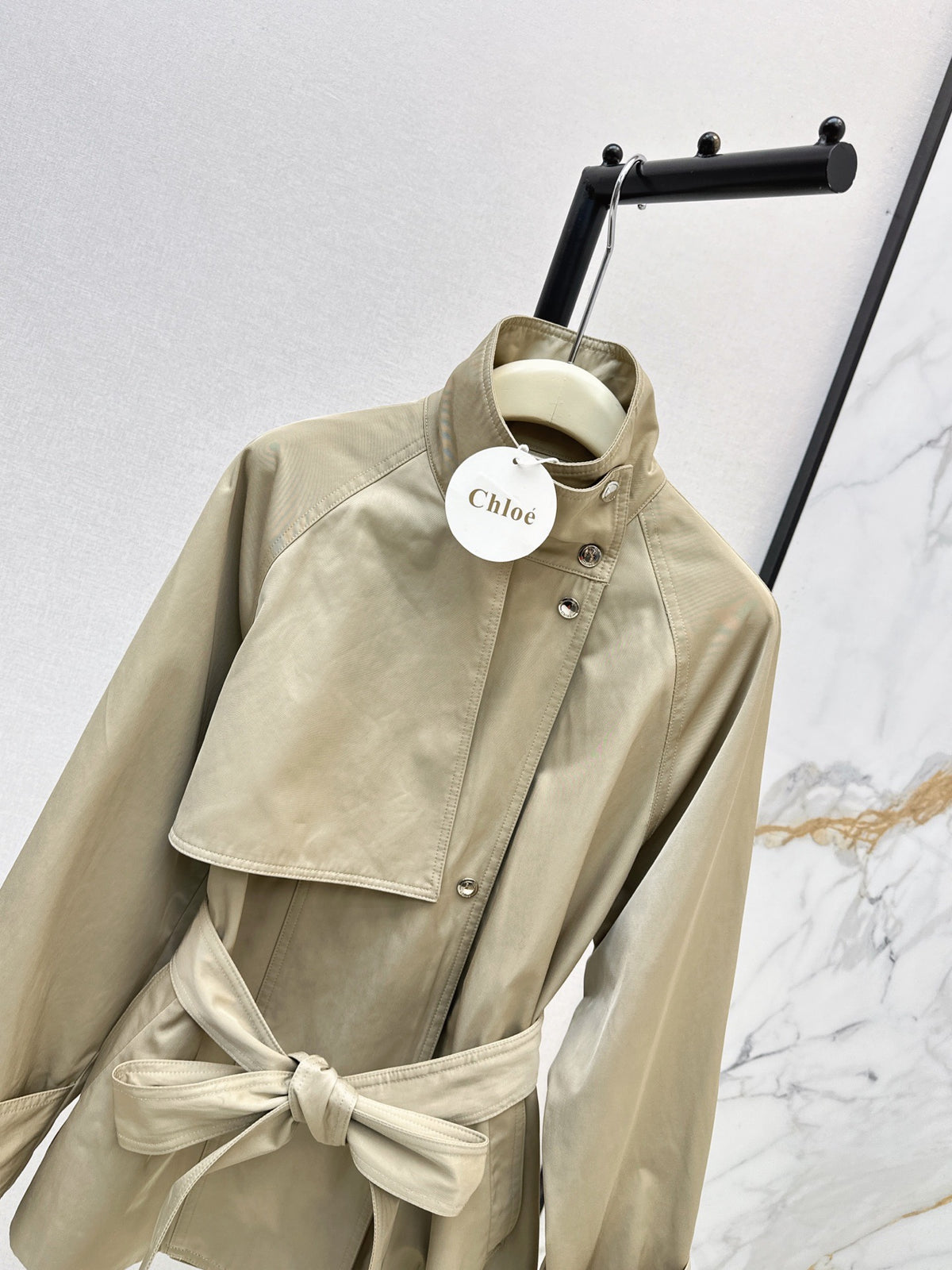 A beige Chloé belted jacket with a high collar and asymmetrical button closure, hanging on a black clothing rack against a light gray and white marble-patterned wall.