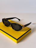 Black cat-eye Fendi sunglasses with gold-tone hardware resting on a bright yellow branded box against a clean off-white background.