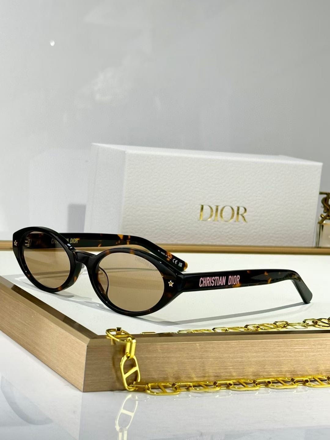 Christian Dior sunglasses with a tortoiseshell pattern, black frames, and gold lenses, resting on a gold chain in front of a white Dior box.