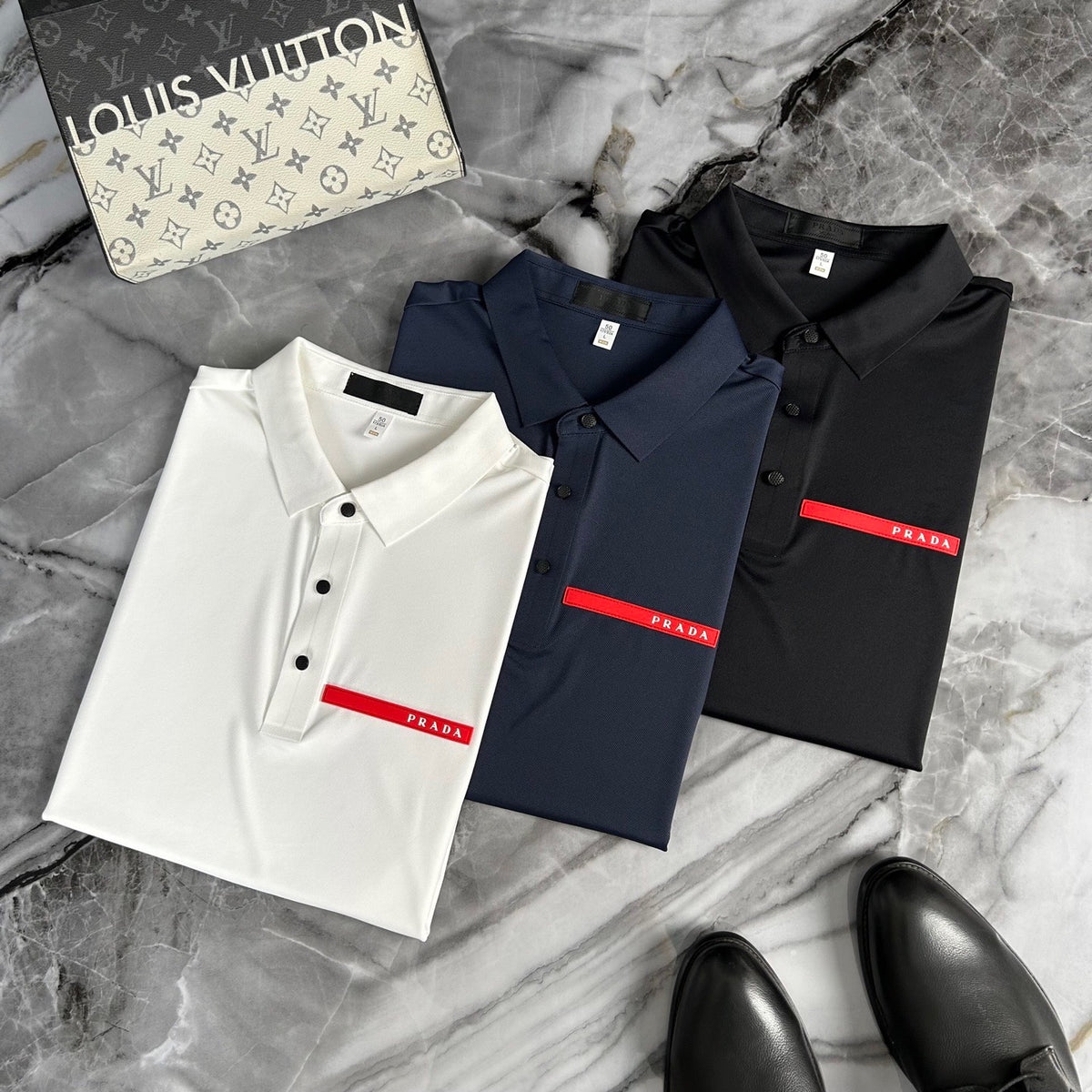 Three folded Prada polo shirts in white, navy blue, and black, each featuring a signature red stripe with the Prada logo on the chest. They are arranged on a gray marble surface next to a Louis Vuitton box and black leather shoes.