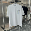 A white Dior short-sleeve t-shirt with a black logo and colorful paint-drip graphic on the chest, hanging on a clear hanger in a retail display.