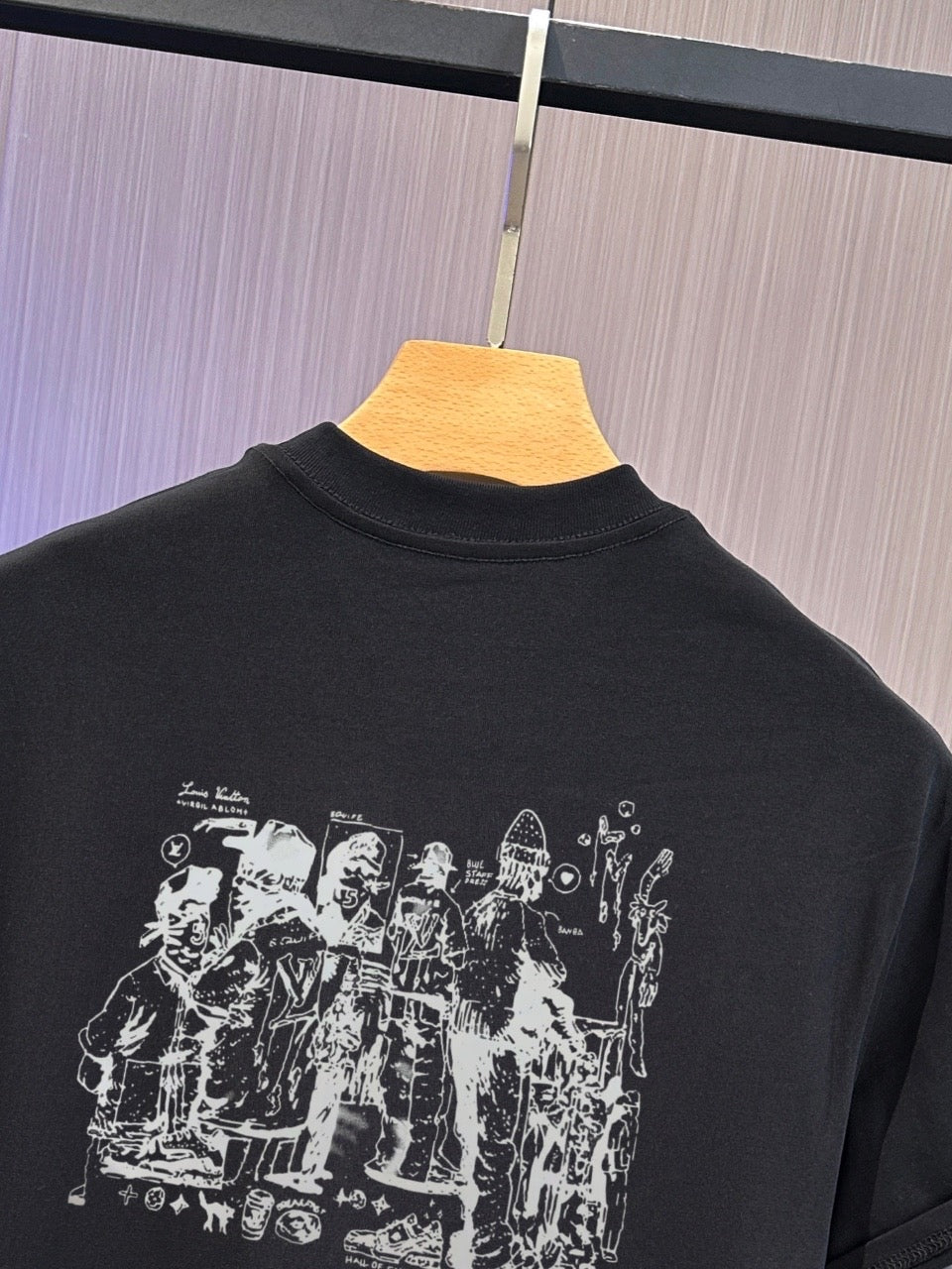 A black short-sleeve t-shirt hanging on a wooden hanger against a light gray paneled wall. The back of the shirt features a large white graphic print depicting a group of historical figures.