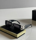 Black Balenciaga square sunglasses with dark lenses and a prominent silver 'BB' logo embellished with clear rhinestones on the side, resting on a black and gold presentation box.