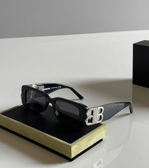 Black Balenciaga square sunglasses with dark lenses and a prominent silver 'BB' logo embellished with clear rhinestones on the side, resting on a black and gold presentation box.