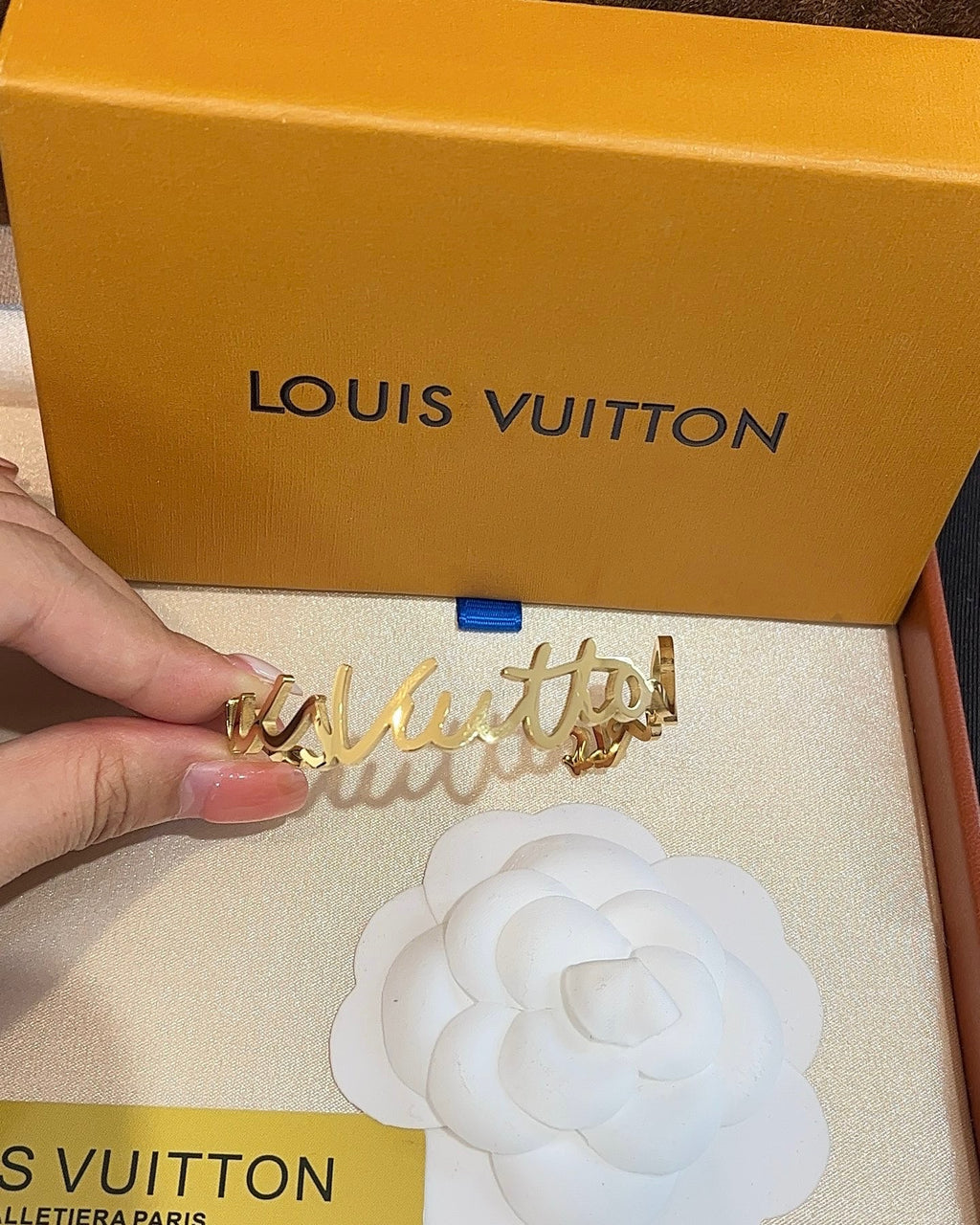 A gold-tone Louis Vuitton script letter accessory held by a hand, with a large orange Louis Vuitton box and a white camellia card in the background.
