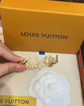 A gold-tone Louis Vuitton script letter accessory held by a hand, with a large orange Louis Vuitton box and a white camellia card in the background.