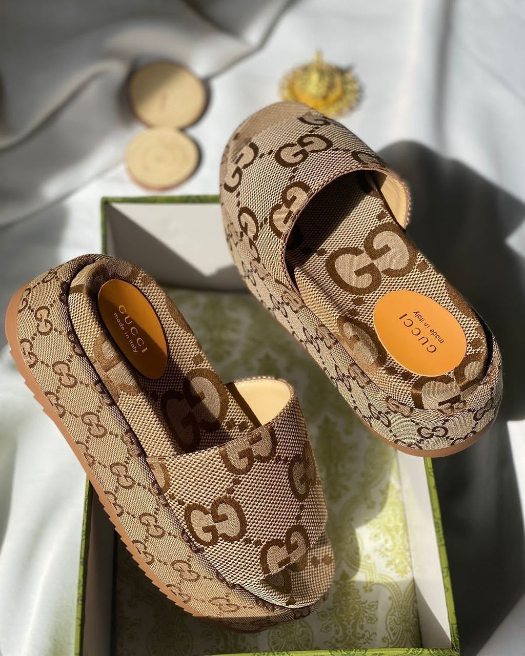 A pair of Gucci slip-on loafers in the signature GG Supreme canvas, displayed on top of their green and white box. The shoes feature the classic brown GG monogram pattern on a beige canvas background, with a tan leather insole and a circular orange brand sticker on the heel.
