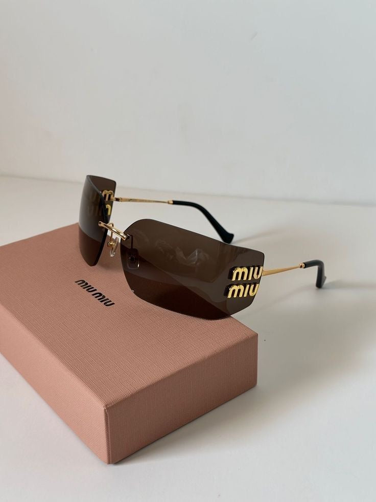 Miu Miu shield sunglasses with dark brown lenses, gold metal frames, and gold 'miu miu' lettering on the temples, resting on a pink Miu Miu box against a white background.