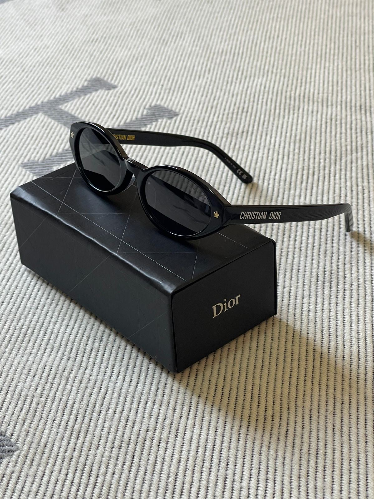 Black Dior sunglasses with dark lenses resting on a black Dior-branded box, placed on a textured beige woven surface. The temple arm shows the Dior name.