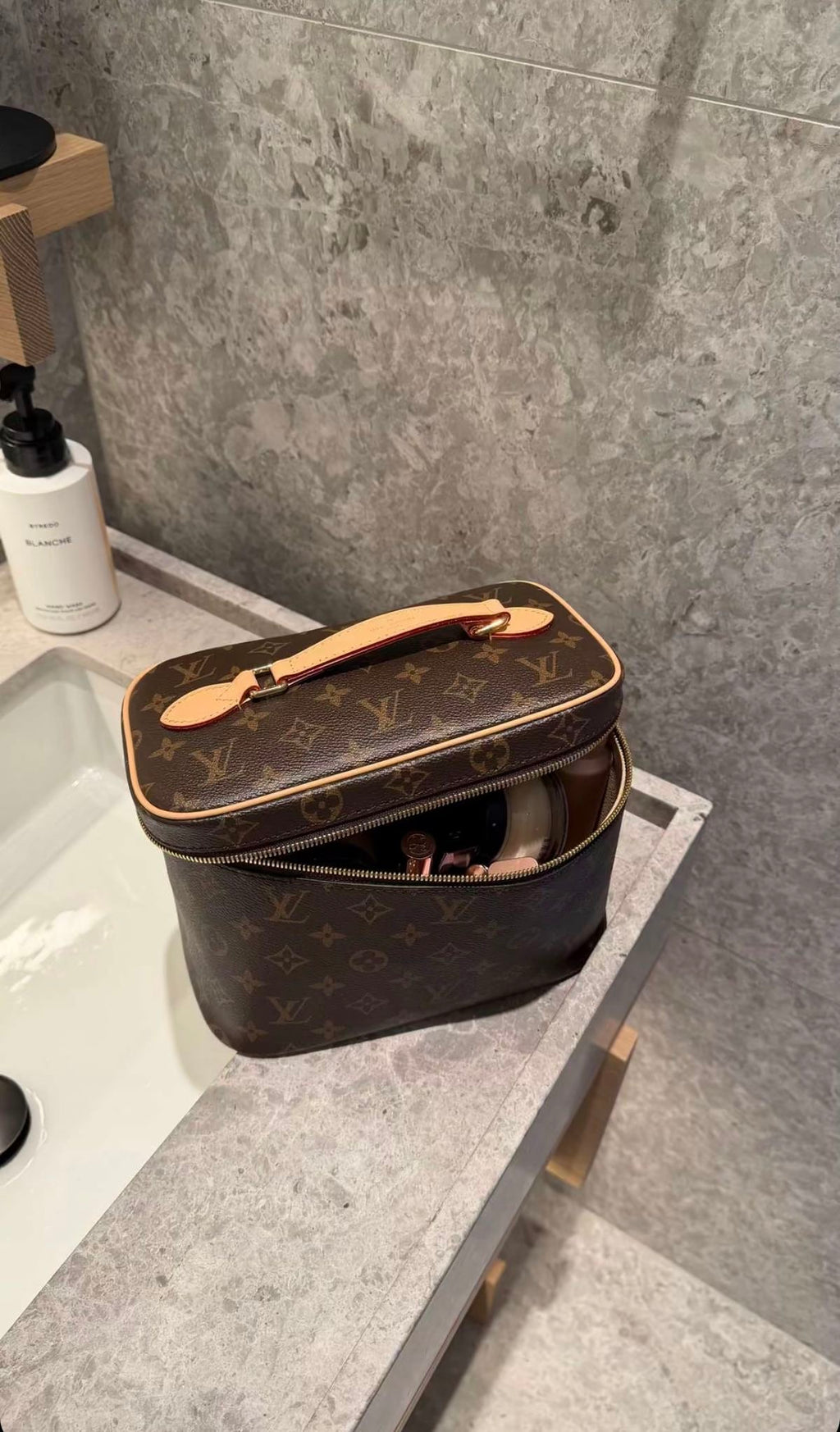 A Louis Vuitton monogram cosmetic bag in brown coated canvas with tan leather trim and handle, sitting open on a gray stone bathroom counter next to a sink.