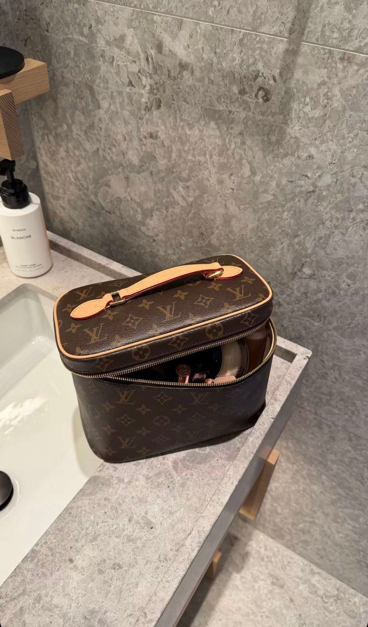 A Louis Vuitton monogram cosmetic bag in brown coated canvas with tan leather trim and handle, sitting open on a gray stone bathroom counter next to a sink.