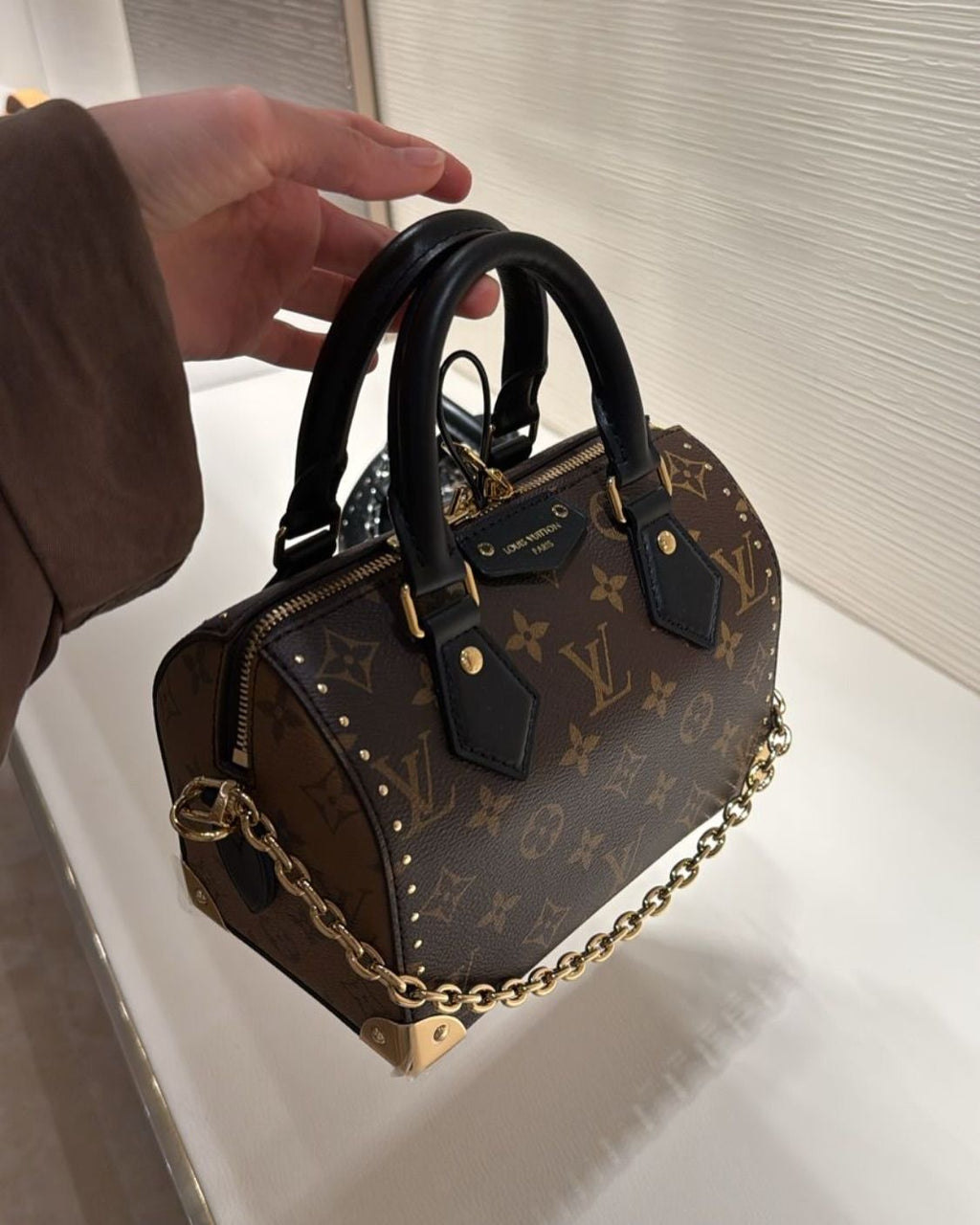 A Louis Vuitton monogram mini top-handle handbag with black leather handles and a gold-tone chain strap, held by a hand against a clean indoor background.