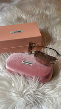 Miu Miu sunglasses with rose gold frames and lenses, displayed next to their signature pink velvet case and peach-colored box on a white faux fur background.