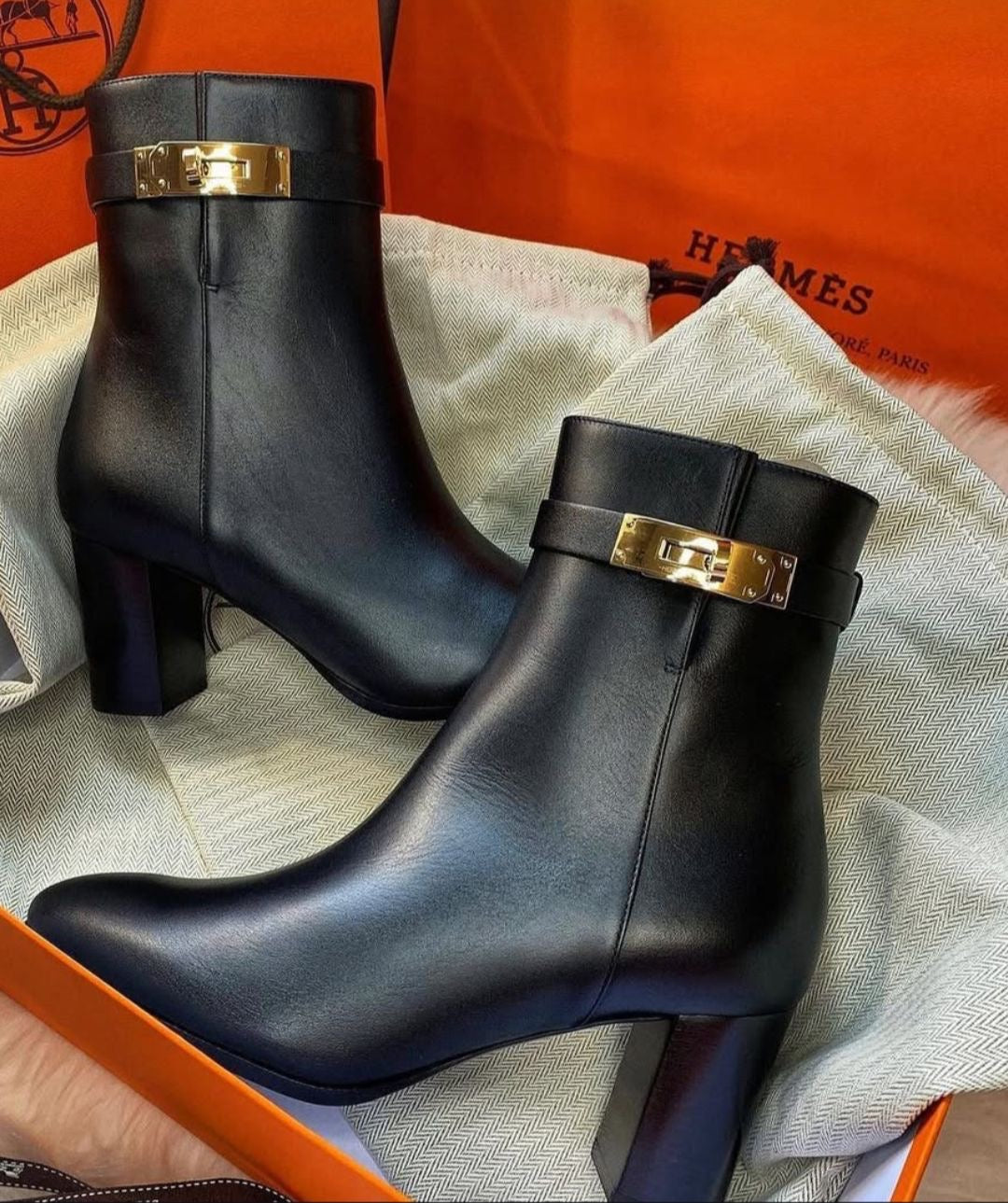 A pair of black leather ankle boots with chunky block heels and gold-tone Hermes hardware, displayed on a light beige dust bag with orange Hermès shoebox packaging visible in the background.