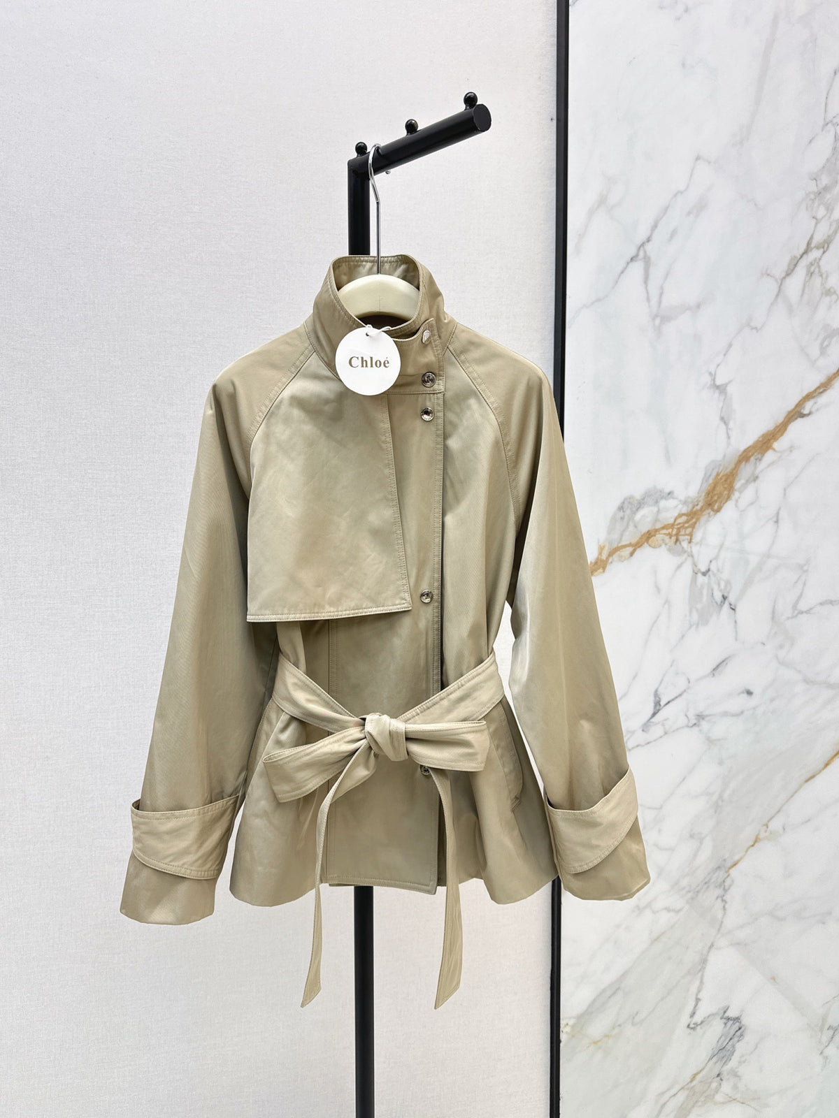 A beige Chloé belted jacket with a high collar and asymmetrical flap detail, displayed on a black hanger stand against a split white and marble background.
