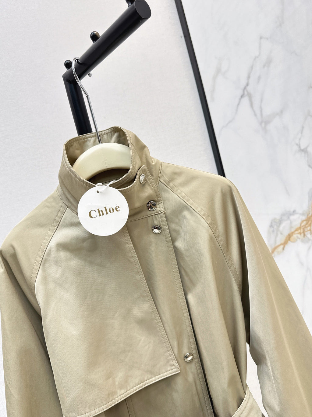 A beige Chloé jacket hanging on a black hanger stand against a white marble-patterned wall. The jacket features a high collar, silver snap buttons, and a white circular Chloé brand tag.