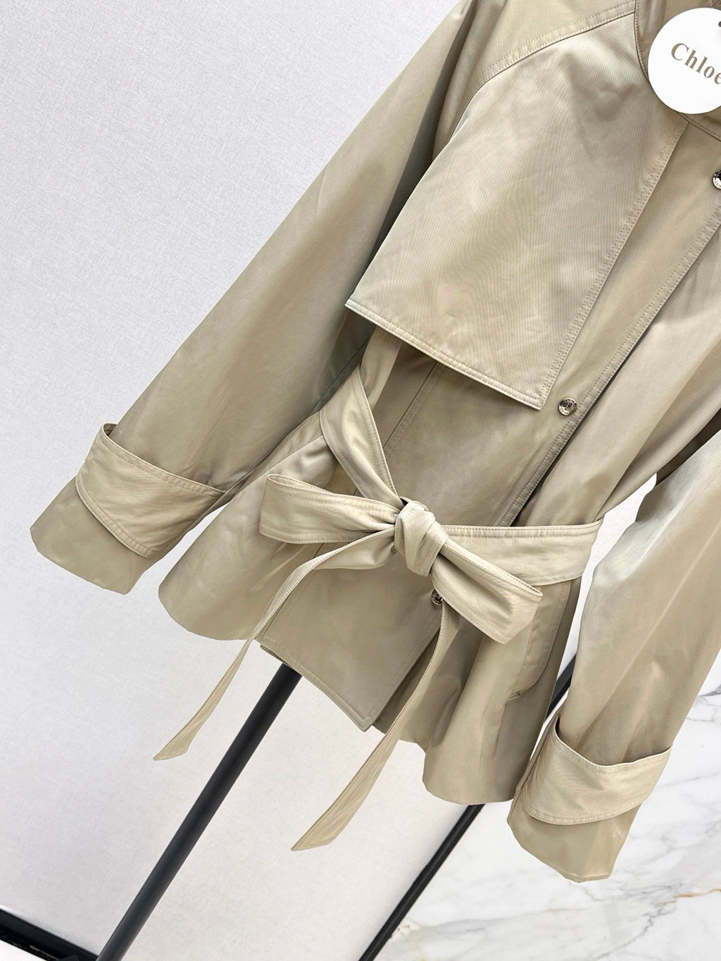 A beige trench coat with a matching waist tie, displayed on a black hanger against a light gray background.