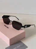 Miu Miu sunglasses with a black geometric frame and dark lenses, resting on a pink Miu Miu box with a gray card in the foreground.