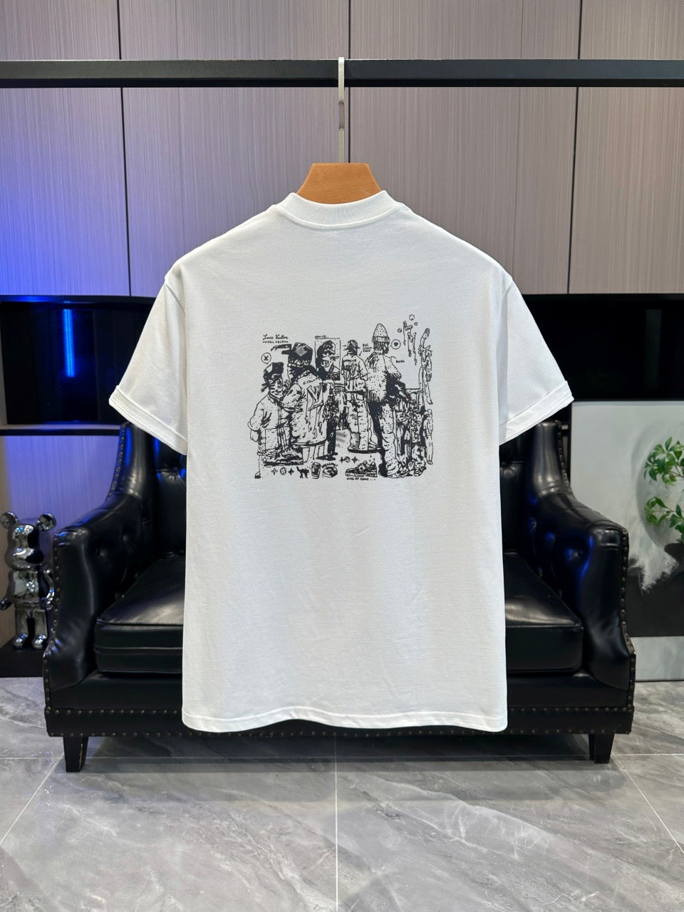 A white short-sleeve t-shirt hanging on a wooden hanger, showing the back with a black graphic print of figures in armor. The shirt is displayed in front of a black leather armchair against a gray paneled wall.