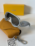 Loewe aviator-style sunglasses with silver frames and dark lenses, displayed with a tan textured leather case, a yellow Loewe box, and a Loewe keychain tag on a white surface.