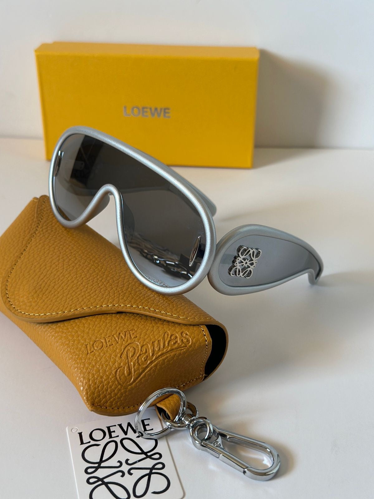 Loewe aviator-style sunglasses with silver frames and dark lenses, displayed with a tan textured leather case, a yellow Loewe box, and a Loewe keychain tag on a white surface.