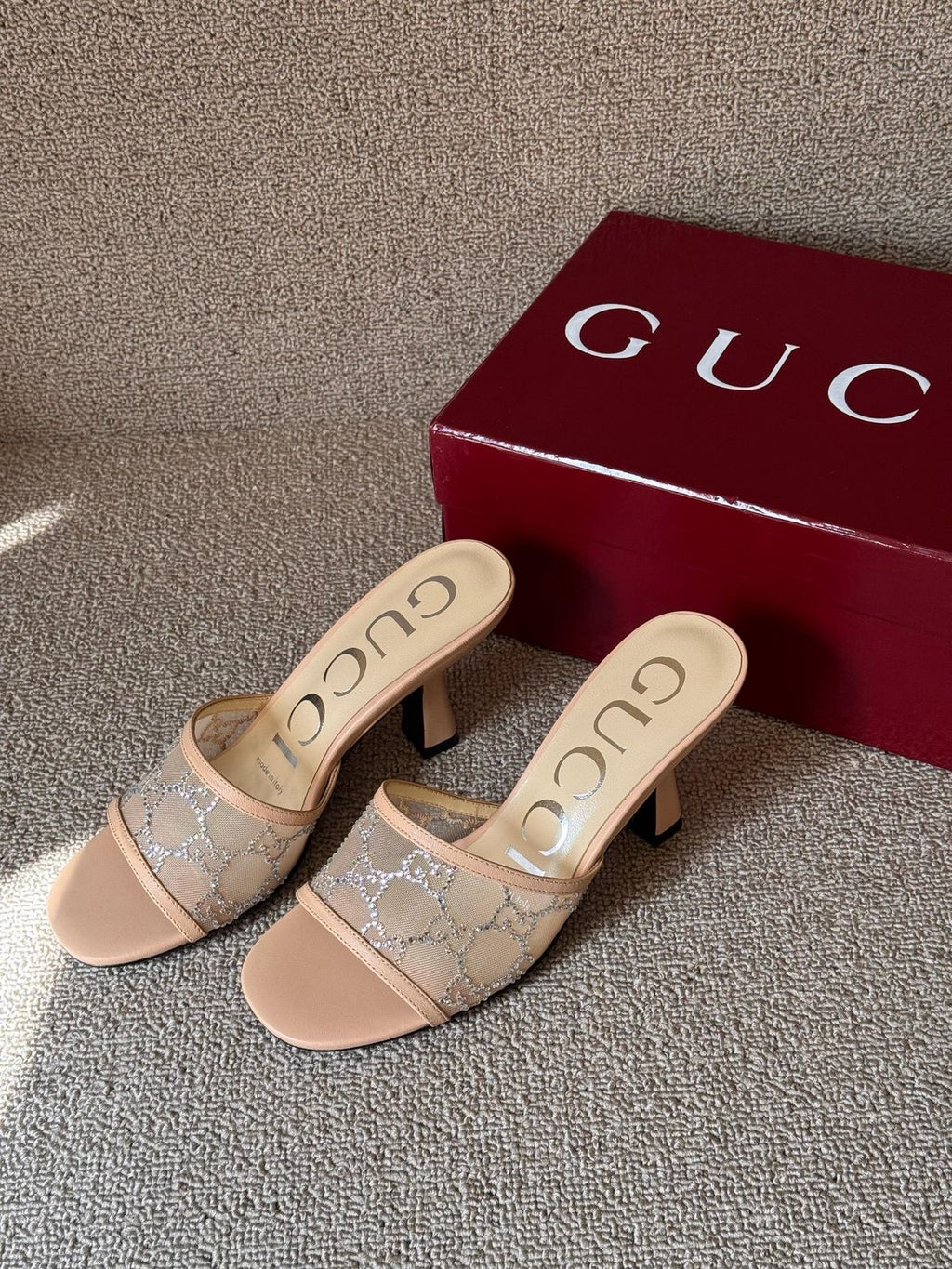 A pair of beige Gucci heeled mule sandals with a tonal GG monogram pattern, displayed next to a dark red Gucci shoebox on a textured gray carpet.