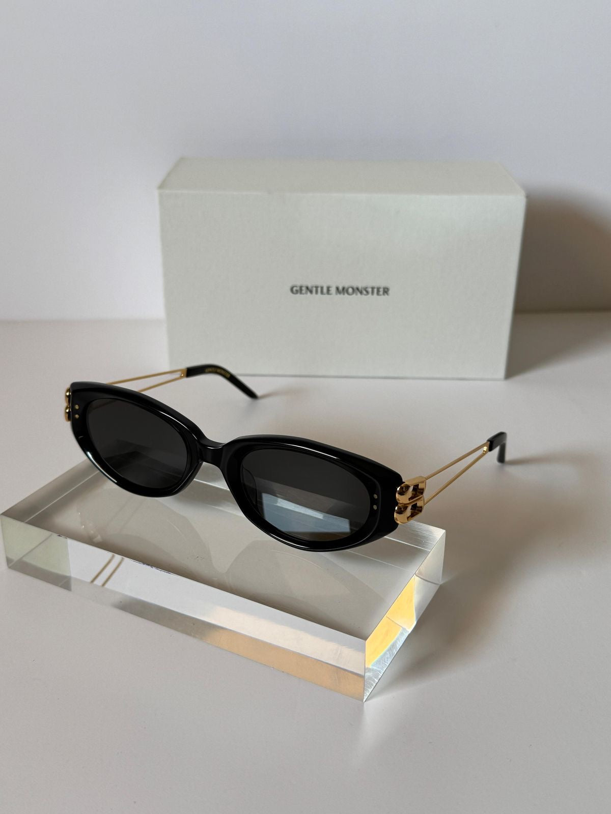 Gentle Monster sunglasses with black acetate frames, gold metal temples, and dark lenses, displayed on a clear acrylic block with their branded white box in the background.