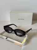 Black oval sunglasses with dark lenses displayed on a clear acrylic block, with a white Gentle Monster box in the background.