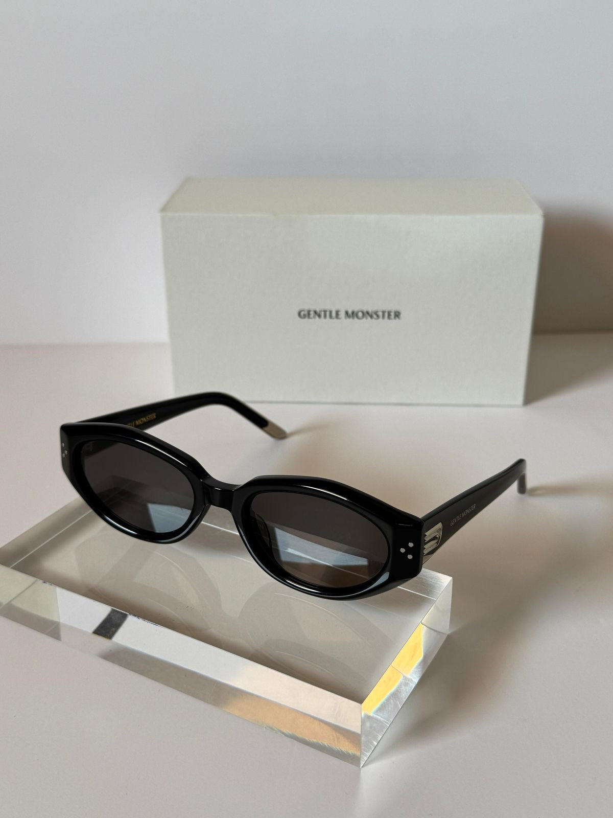 Black oval sunglasses with dark lenses displayed on a clear acrylic block, with a white Gentle Monster box in the background.