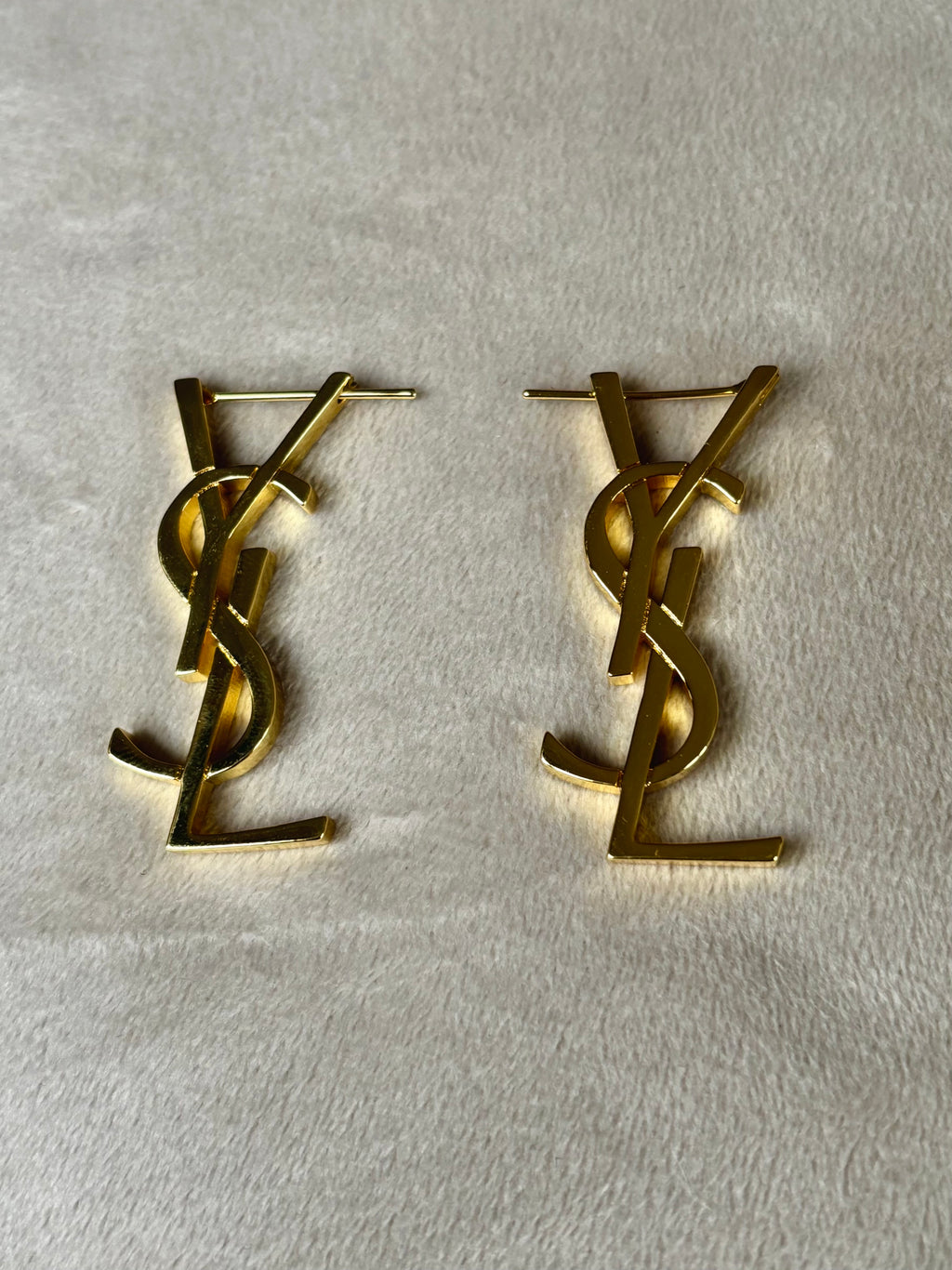 A pair of gold-tone metal stud earrings featuring the interlocking YSL logo, displayed on a textured off-white fabric background.