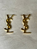 A pair of gold-tone metal stud earrings featuring the interlocking YSL logo, displayed on a textured off-white fabric background.