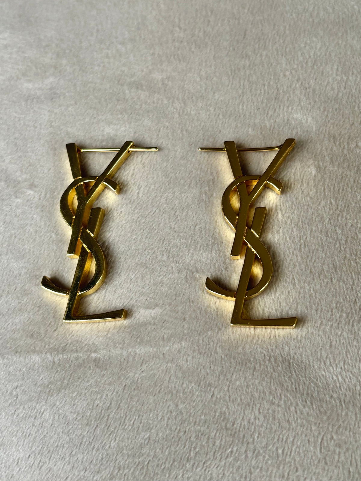 A pair of gold-tone metal stud earrings featuring the interlocking YSL logo, displayed on a textured off-white fabric background.