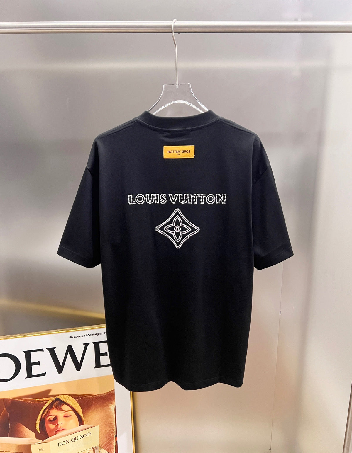 A black Louis Vuitton short-sleeve t-shirt hanging on a clear plastic hanger against a metallic wall. The back of the shirt features the white 'LOUIS VUITTON' text and a white outlined diamond monogram logo.