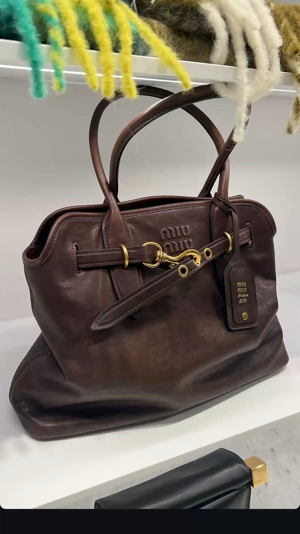 A dark brown leather Miu Miu handbag with gold-tone hardware, displayed on a white shelf with a colorful fuzzy textile hanging above.