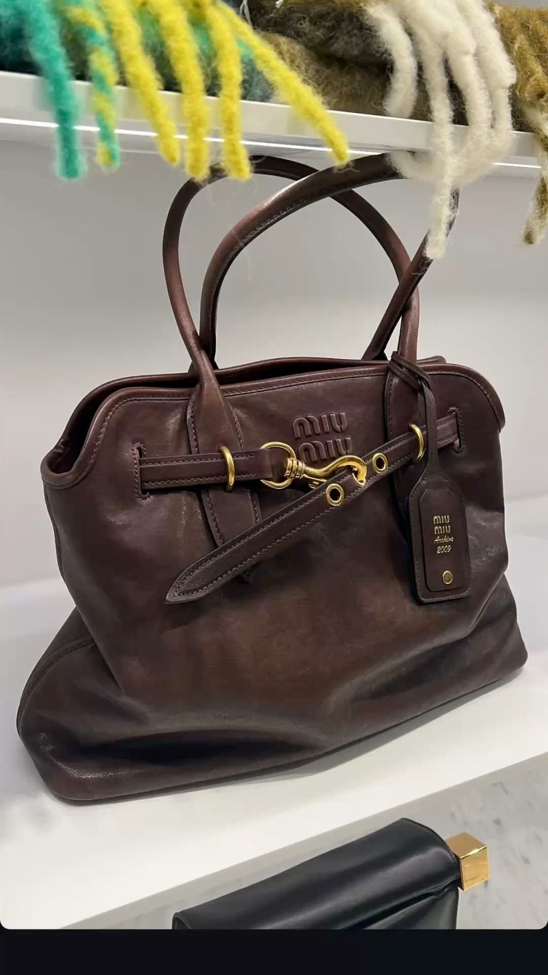 A dark brown leather Miu Miu handbag with gold-tone hardware, displayed on a white shelf with a colorful fuzzy textile hanging above.